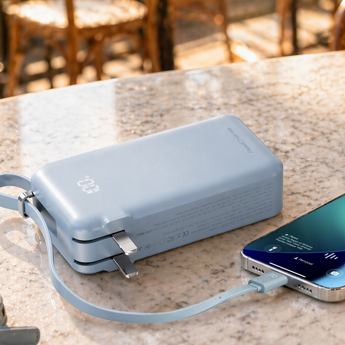 ReelCharge 45W 10,000 mAh Power Bank in Soft Blue charging a phone with its integrated cable on a cafe table